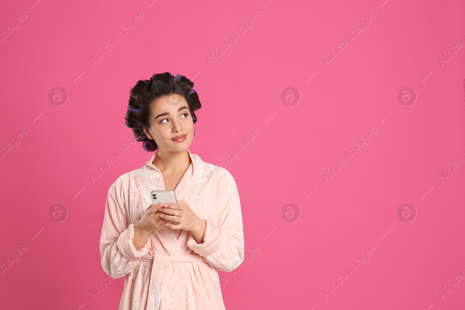 Photo of Happy young woman in bathrobe with hair curlers using smartphone on pink background, space for text