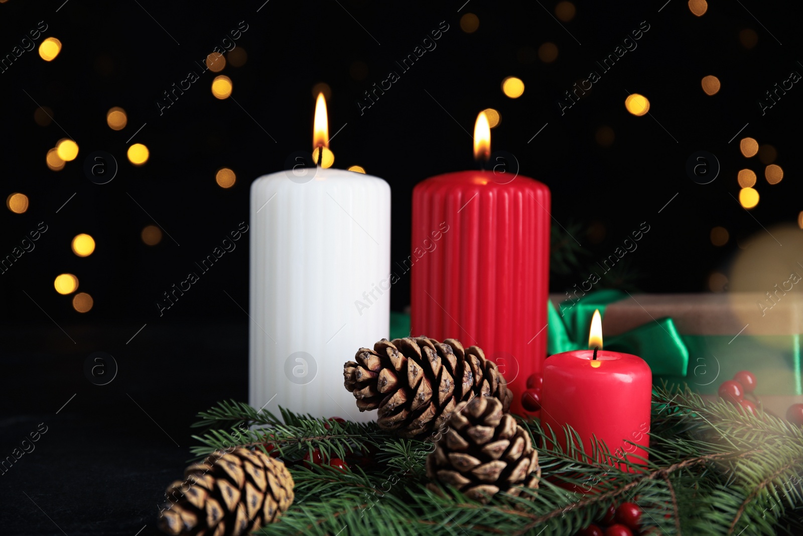 Photo of Burning candles, gift box, fir branches and cones on dark table against blurred Christmas lights