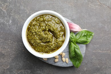 Photo of Tasty pesto sauce in bowl, basil, pine nuts and garlic on grey table, top view