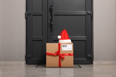 Photo of Christmas gift box and Santa hat near door indoors. Sending present by mail