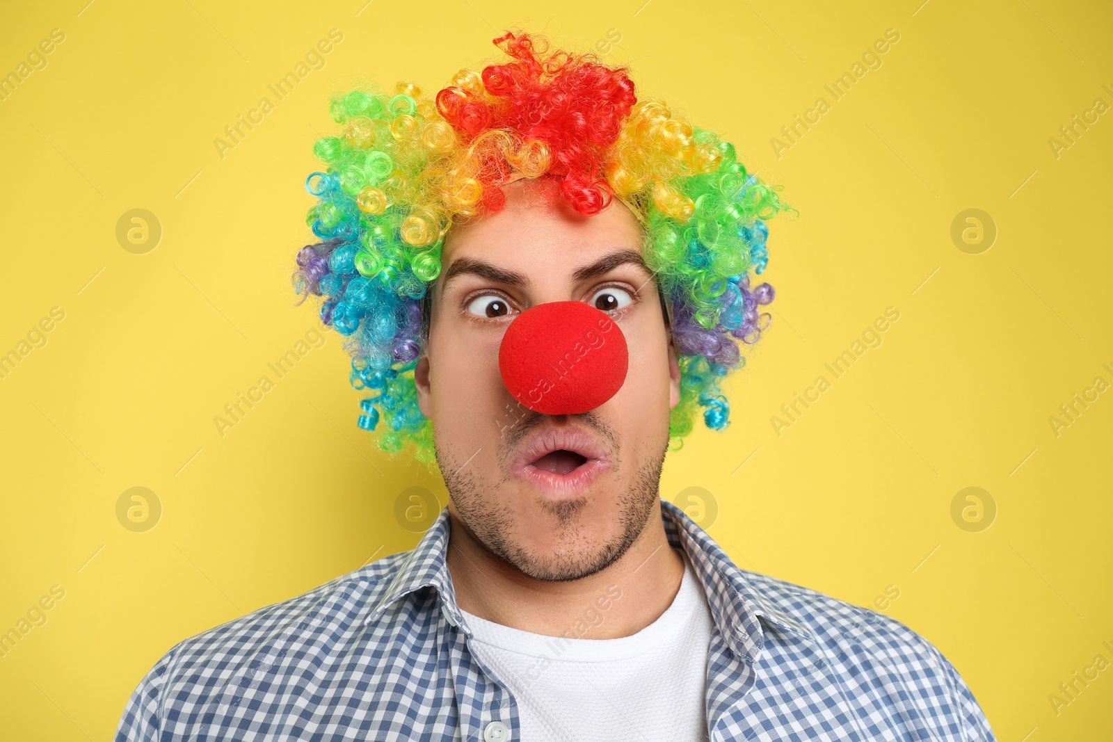 Photo of Funny man with clown nose and rainbow wig on yellow background. April fool's day