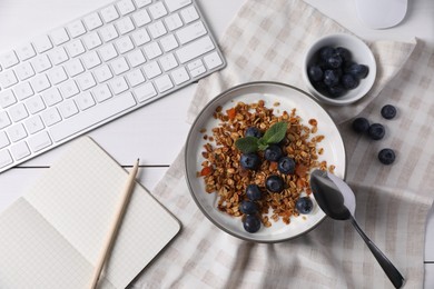 Photo of Delicious granola with blueberries in bowl, stationery and computer keyboard on white wooden table, flat lay