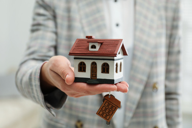 Photo of Real estate agent holding house model and key indoors, closeup