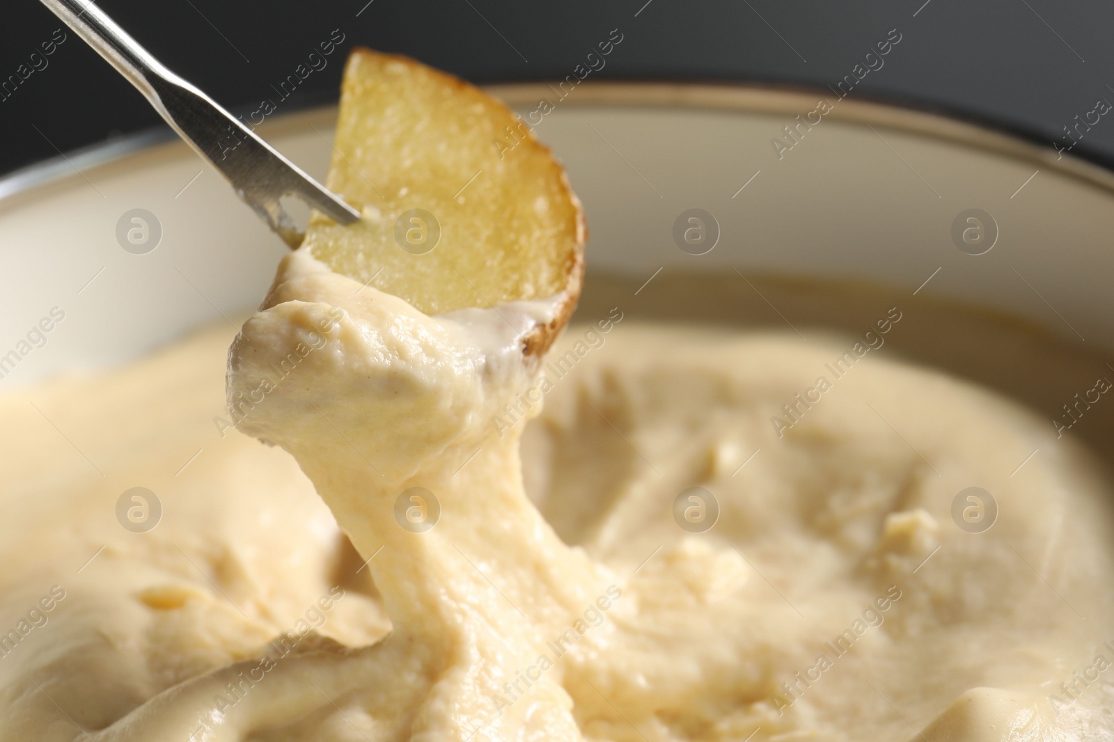 Photo of Dipping piece of potato into fondue pot with melted cheese on grey background, closeup