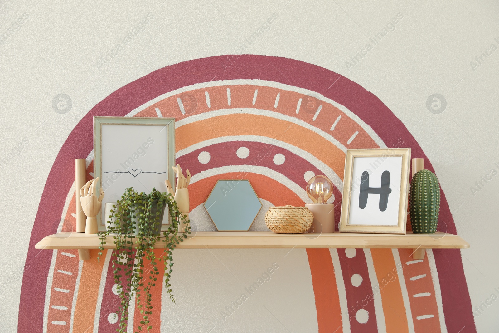 Photo of Stylish decor elements on wooden shelf near white wall with painted rainbow indoors. Interior design