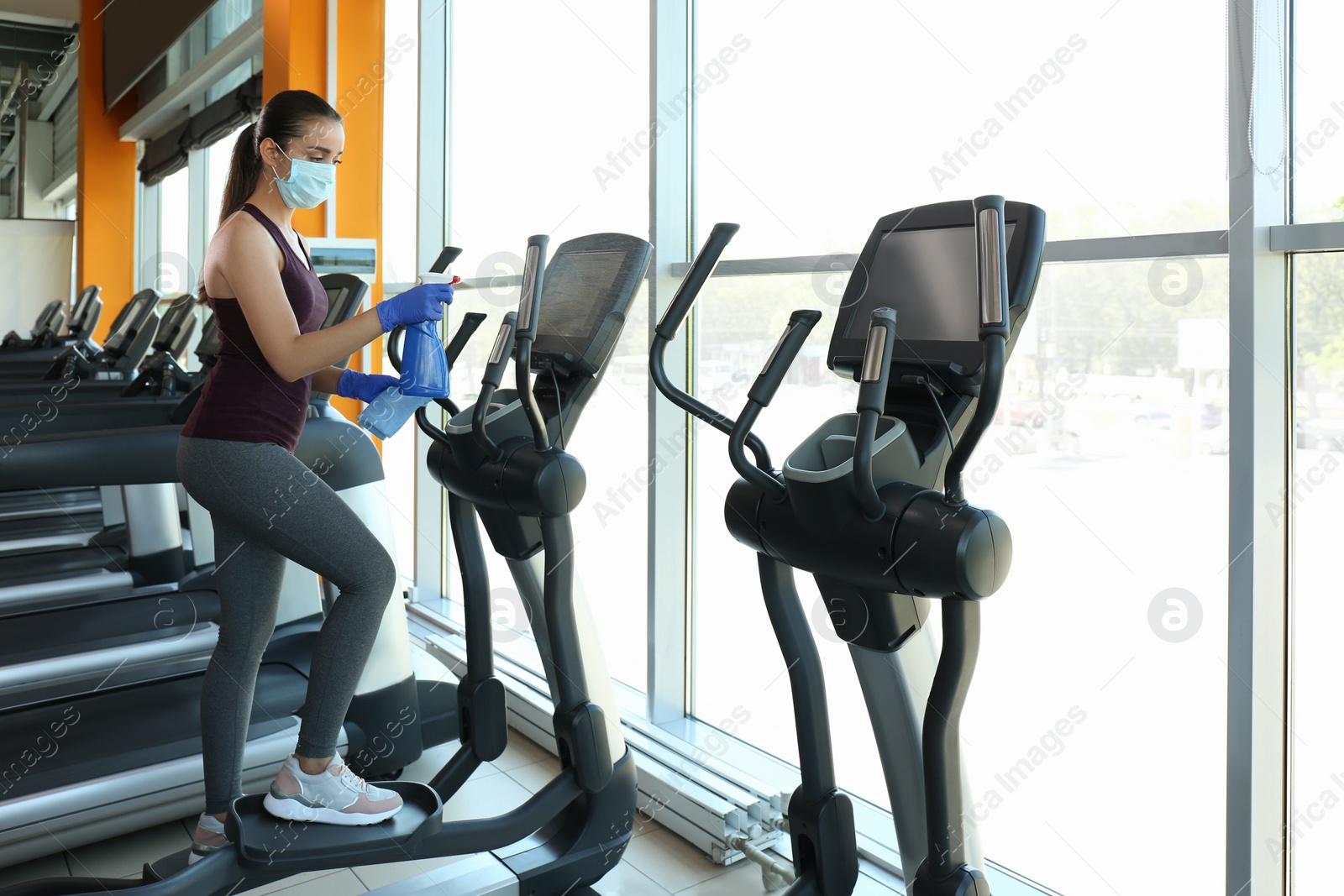 Photo of Woman cleaning exercise equipment with disinfectant spray and cloth in gym