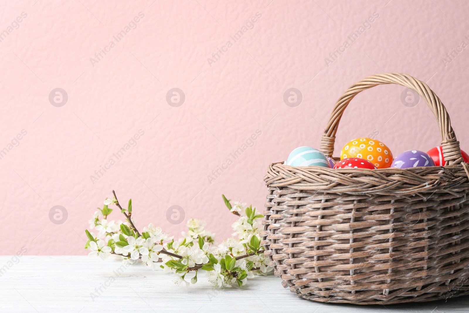 Photo of Painted Easter eggs in wicker basket and blossoming branches on table against color background. Space for text