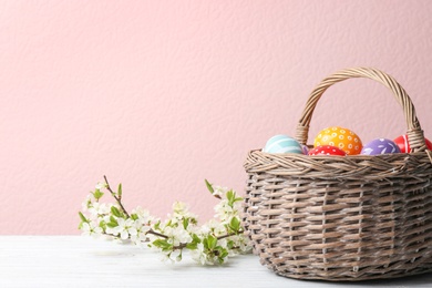 Photo of Painted Easter eggs in wicker basket and blossoming branches on table against color background. Space for text