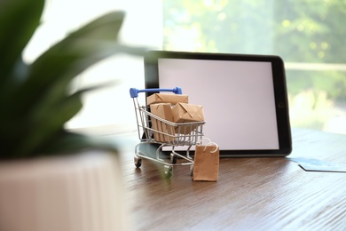 Photo of Internet shopping. Small cart with boxes and bag near modern tablet on wooden table indoors