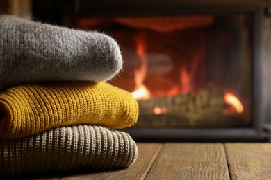 Stack of sweaters on wooden table near fireplace at home, closeup. Space for text