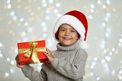 Photo of Happy little child in Santa hat with gift box against blurred festive lights. Christmas celebration
