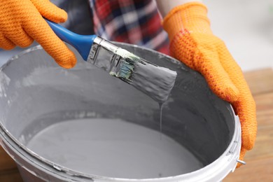 Photo of Woman dipping brush into bucket of grey paint at wooden table, closeup