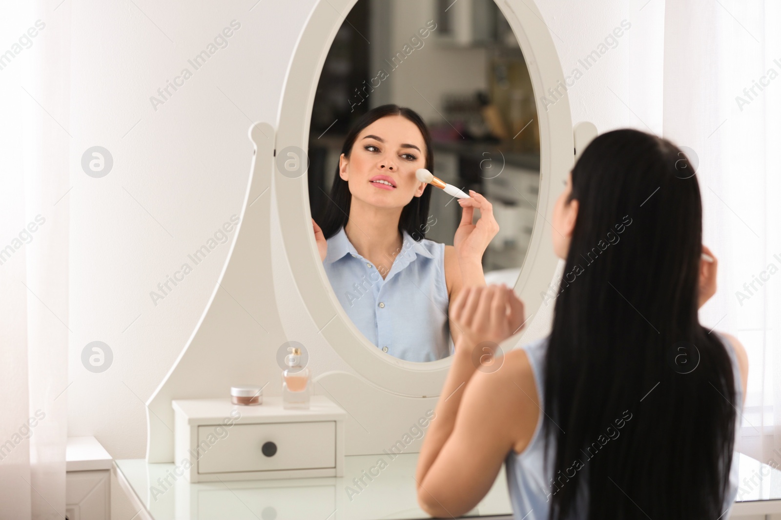 Photo of Beautiful young woman applying makeup near mirror indoors