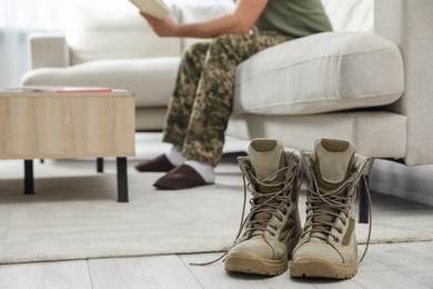 Photo of Soldier with magazine on sofa in living room, focus on pair of combat boots. Military service