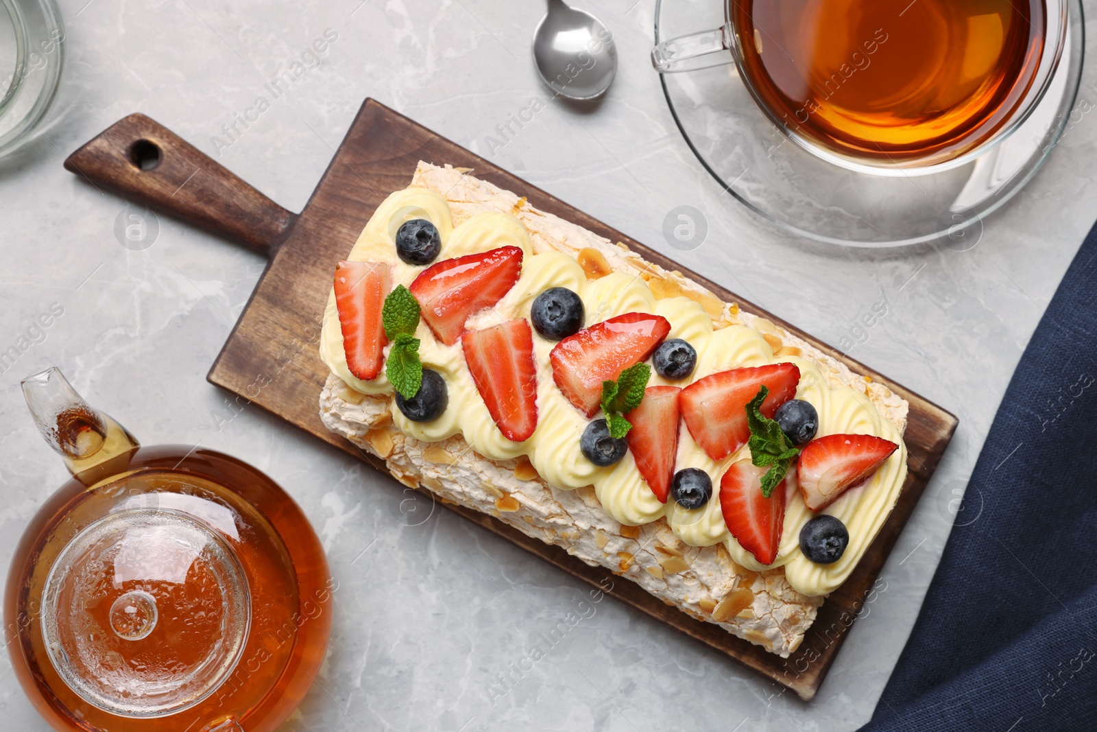 Photo of Tasty meringue roll with cream, strawberry, blueberry, mint and aromatic tea on light grey marble table, flat lay