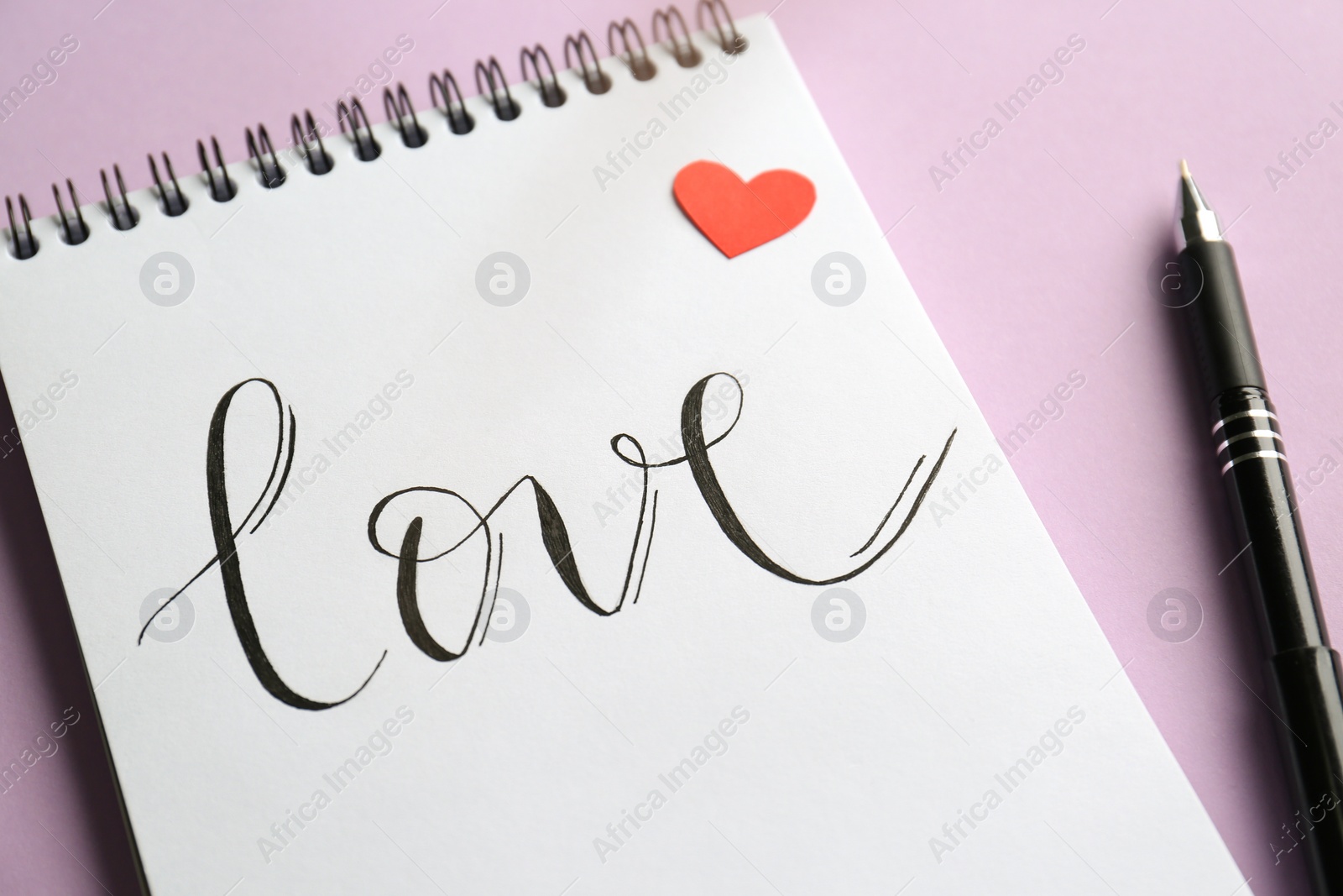 Photo of Notebook with handwritten word Love, red paper heart and pen on violet background, closeup