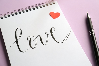 Photo of Notebook with handwritten word Love, red paper heart and pen on violet background, closeup