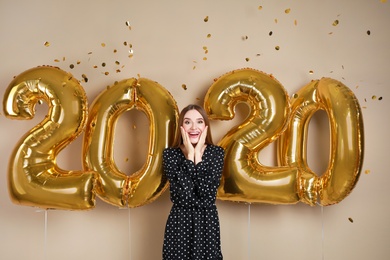 Excited young woman near golden 2020 balloons on beige background. New Year celebration