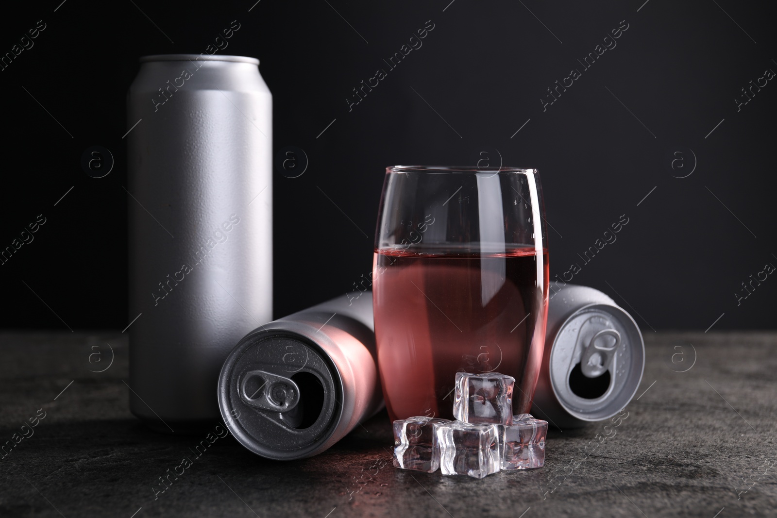 Photo of Energy drink in glass, aluminium cans and ice cubes on grey table