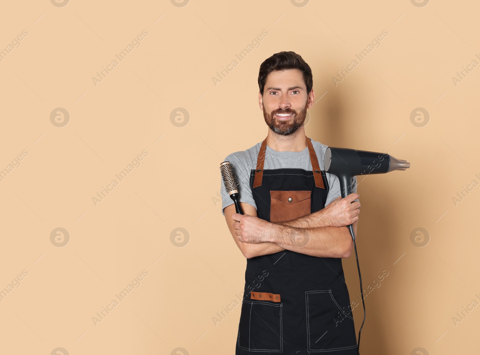 Photo of Smiling hairdresser in apron holding dryer and brush on light brown background, space for text