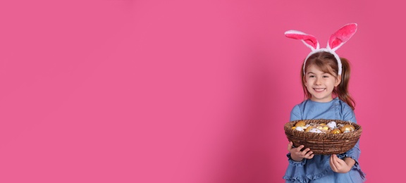 Happy little girl with bunny ears holding wicker basket full of Easter eggs on pink background. Space for text