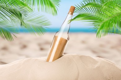 Image of Corked glass bottle with rolled paper note on sandy beach with palms near ocean
