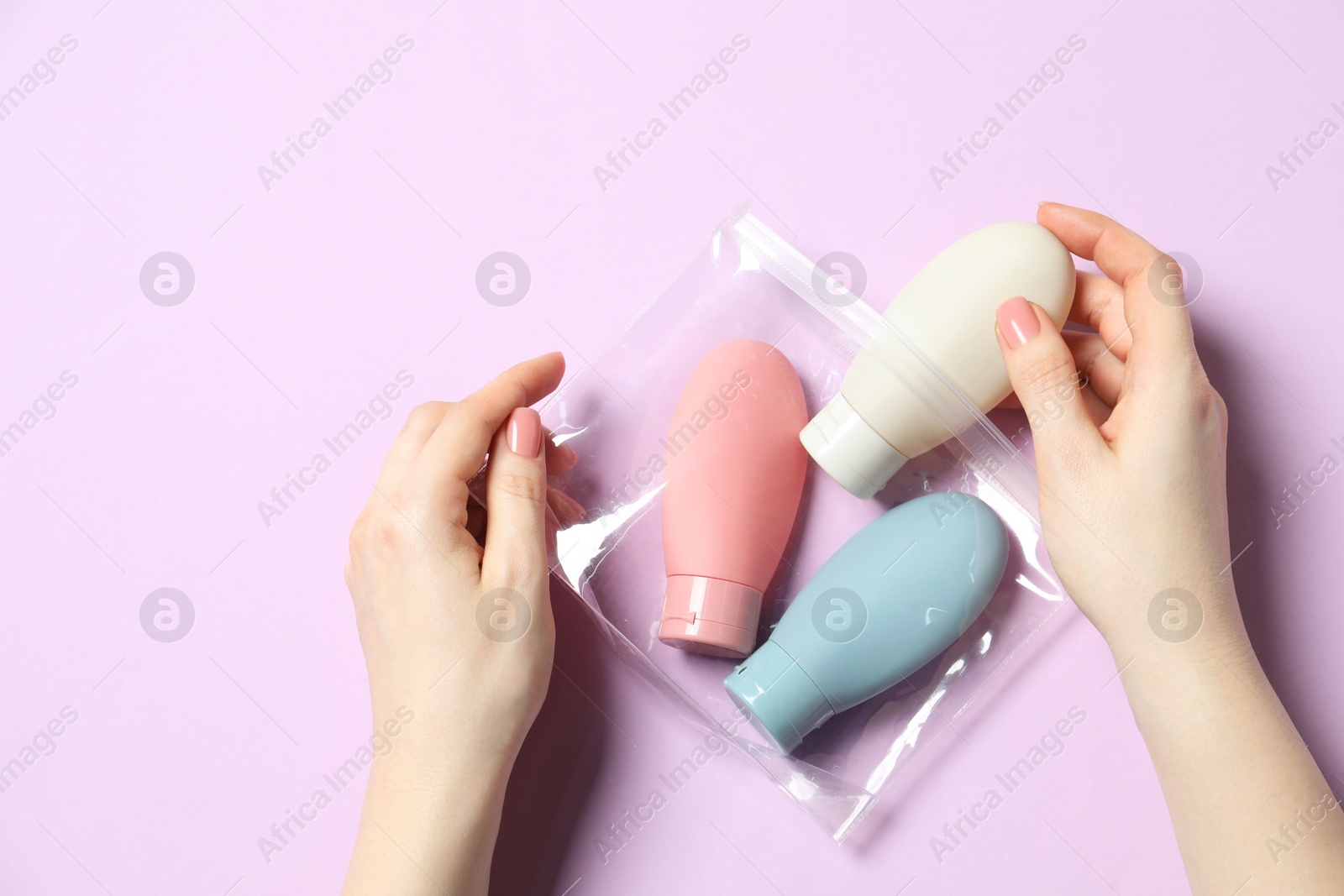 Photo of Cosmetic travel kit. Woman putting small bottle with personal care product into plastic bag against violet background, top view