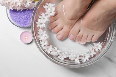 Photo of Woman soaking her feet in bowl with water and flowers on white marble table, top view. Pedicure procedure