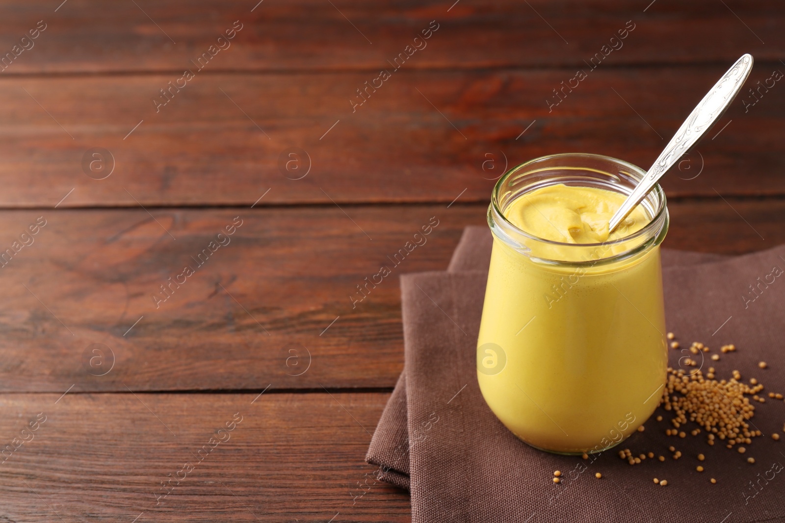 Photo of Tasty mustard sauce and spoon in glass jar with dry seeds on wooden table, space for text