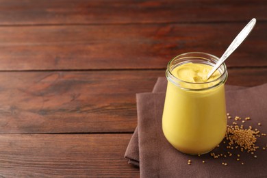 Tasty mustard sauce and spoon in glass jar with dry seeds on wooden table, space for text