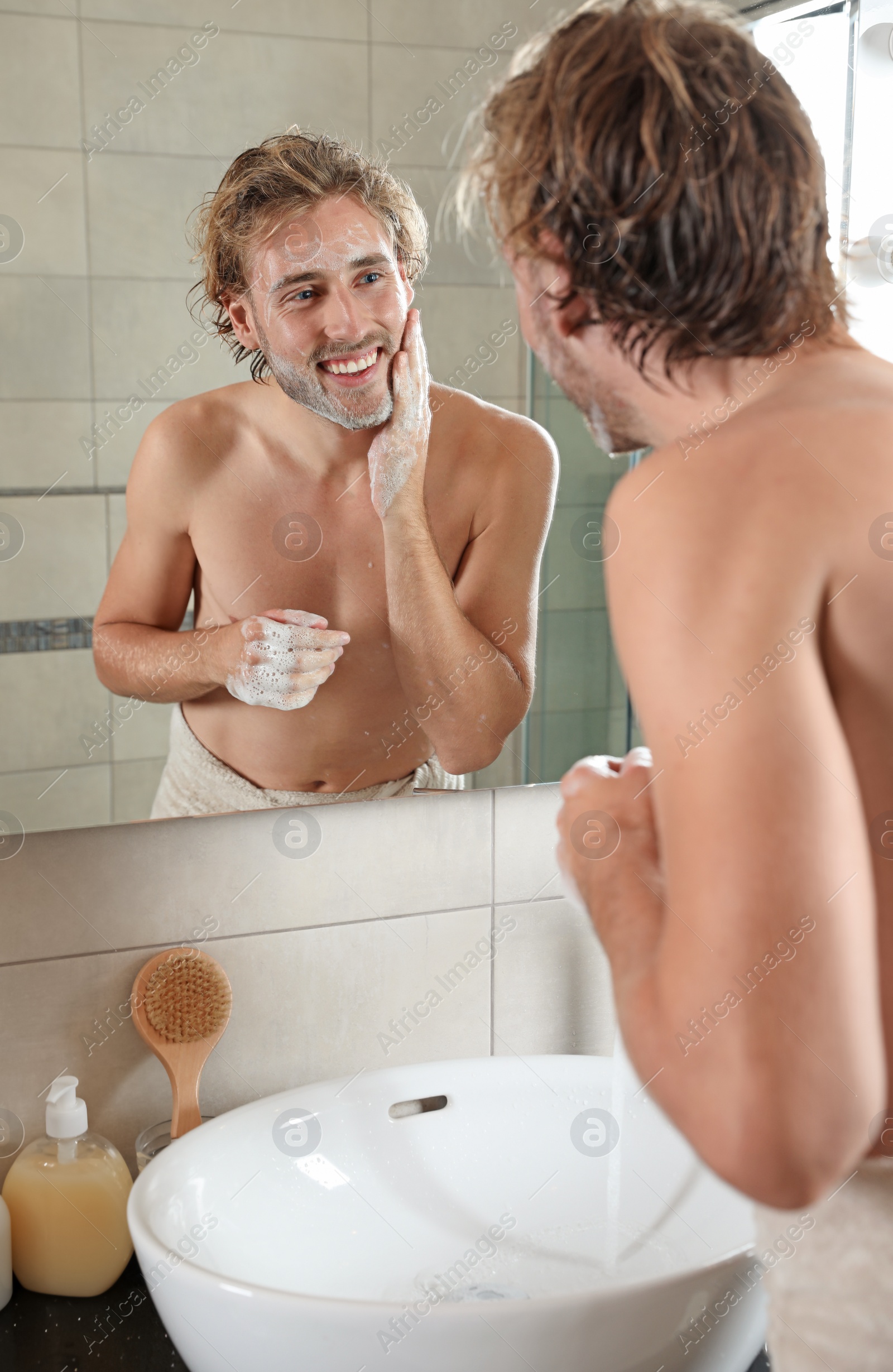 Photo of Young man washing face with soap near mirror in bathroom