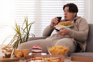 Photo of Overweight man with plate of burgers and French fries watching TV on sofa at home
