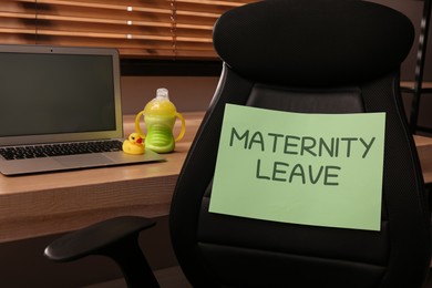 Baby accessories and laptop on wooden table near chair with note Maternity Leave in office