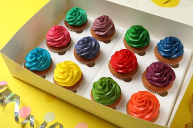 Box with different cupcakes and confetti on yellow background, closeup
