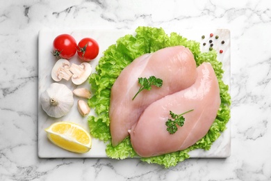 Board with raw chicken breasts and ingredients on marble background, top view