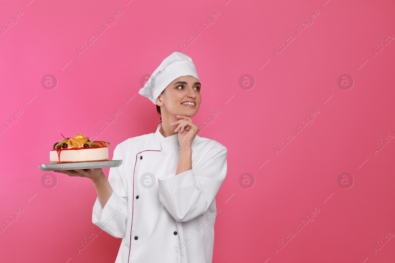 Photo of Happy professional confectioner in uniform holding delicious cake on pink background. Space for text