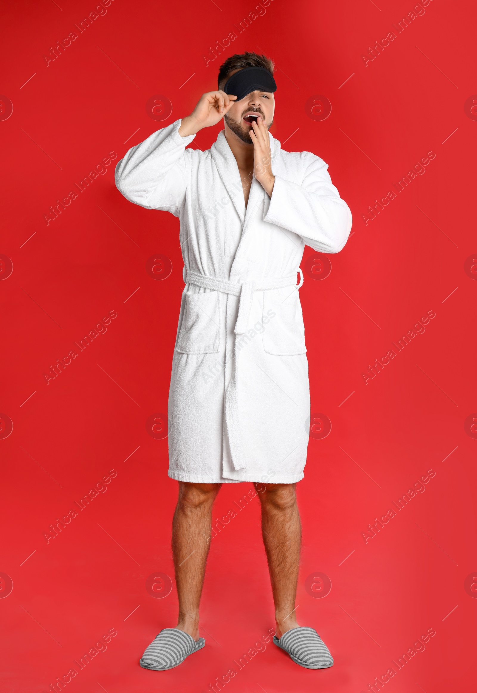 Photo of Young man in bathrobe and eye sleeping mask yawning on red background