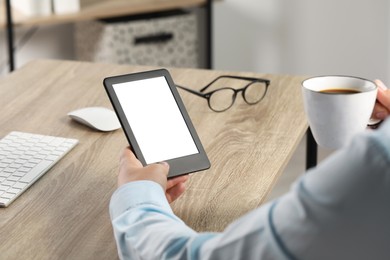 Man with cup of coffee using e-book reader at wooden table indoors, closeup