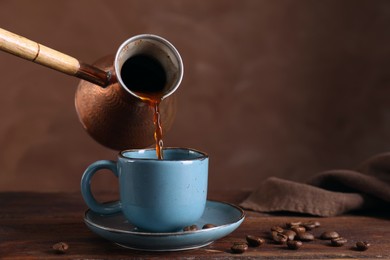 Turkish coffee. Pouring brewed beverage from cezve into cup at wooden table against brown background, space for text