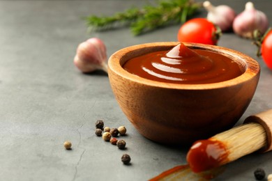 Photo of Tasty barbeque sauce in bowl, brush and peppercorns on grey textured table, closeup. Space for text