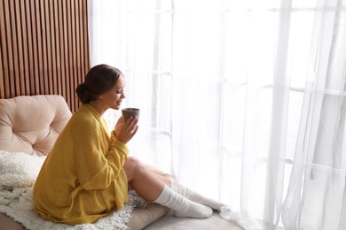 Photo of Beautiful young woman with cup of drink near window at home, space for text. Winter atmosphere