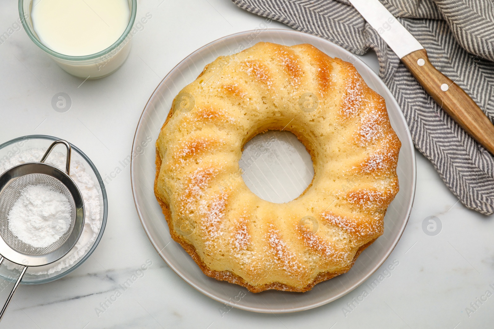 Photo of Delicious sponge cake with powdered sugar, glass of milk and knife on white table, flat lay