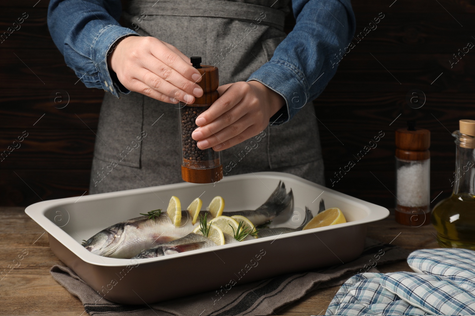 Photo of Woman grinding pepper onto raw sea bass fish with lemon and rosemary at wooden table, closeup