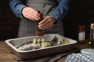 Photo of Woman grinding pepper onto raw sea bass fish with lemon and rosemary at wooden table, closeup