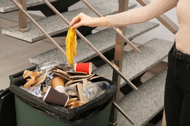 Young woman throwing banana peel in trash bin indoors, closeup. Waste recycling