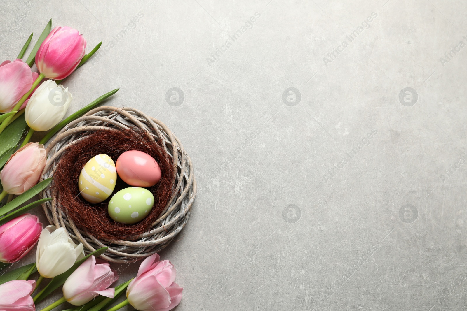 Photo of Flat lay composition with painted Easter eggs in wicker nest and tulips on color background, space for text