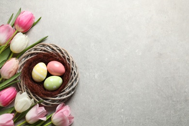 Photo of Flat lay composition with painted Easter eggs in wicker nest and tulips on color background, space for text