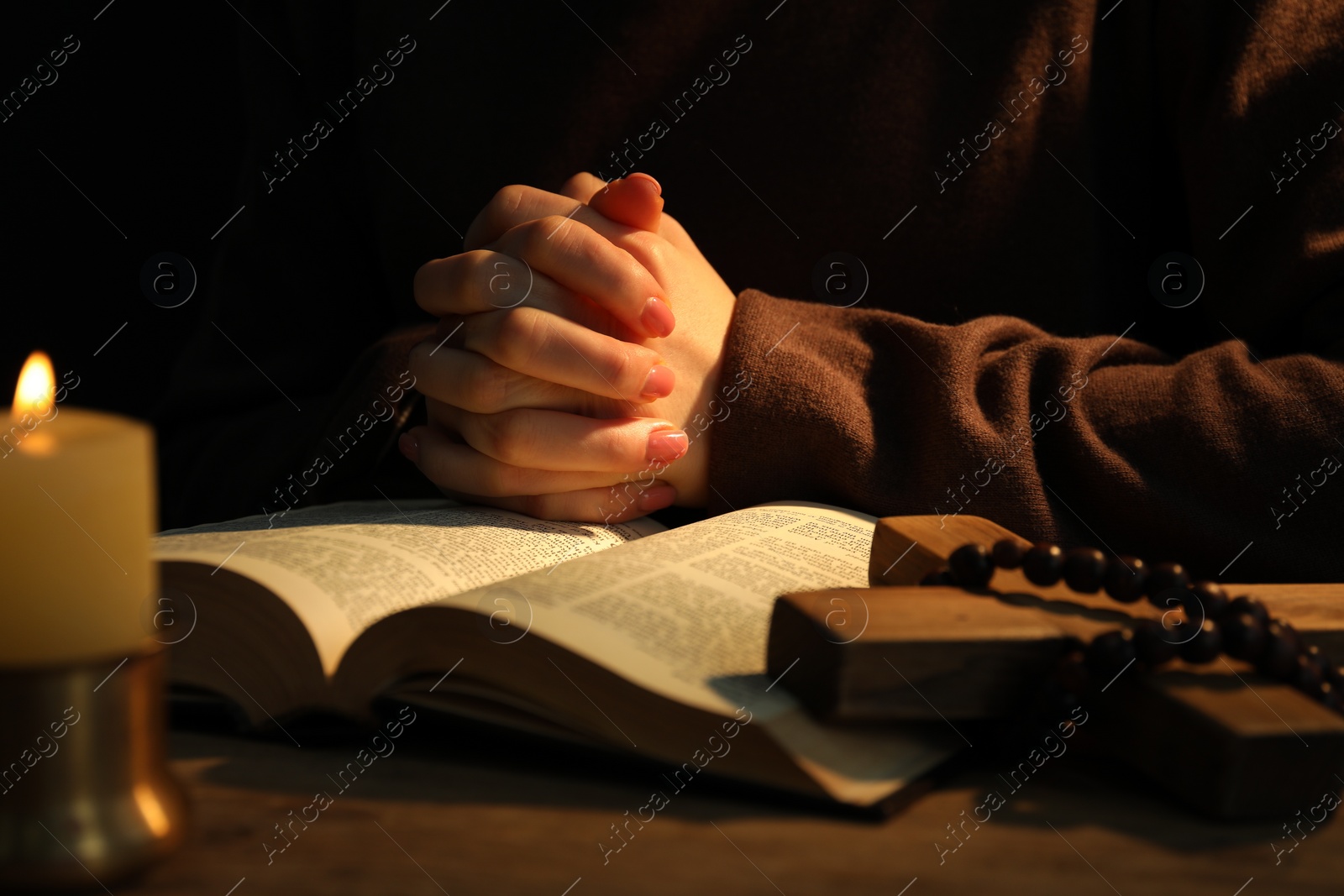 Photo of Woman praying at table with burning candle and Bible, closeup