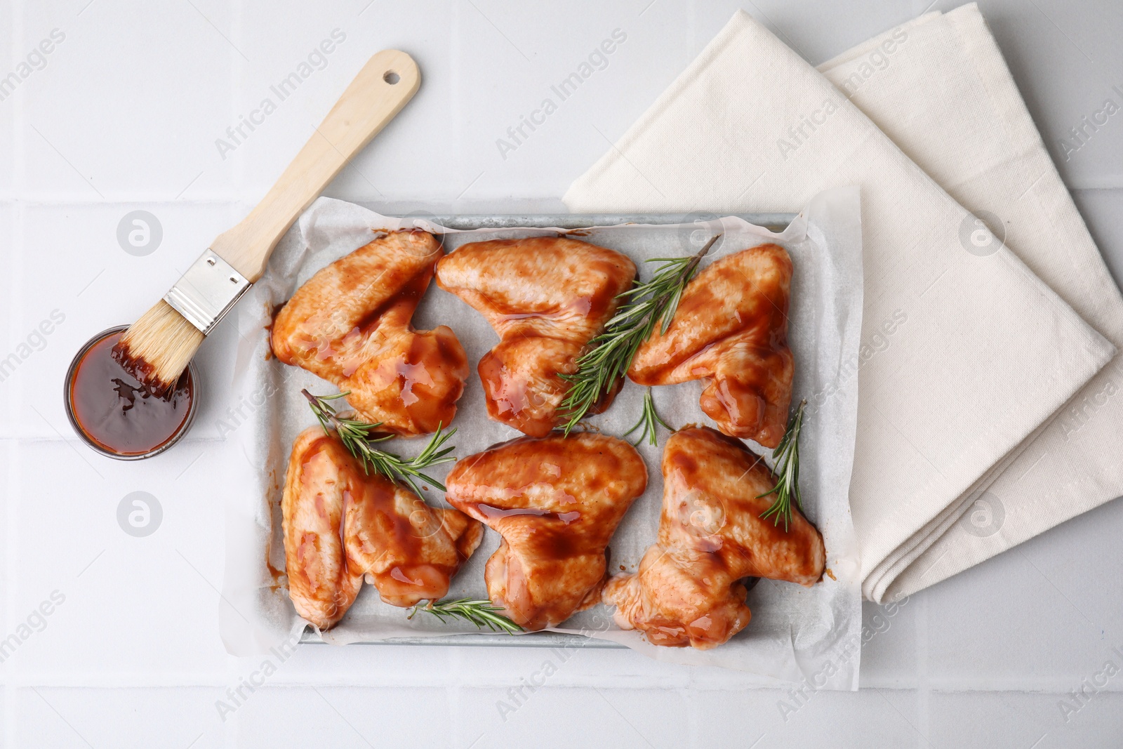 Photo of Raw chicken wings, rosemary, marinade and basting brush on light tiled table, flat lay