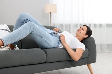 Photo of Young woman struggling to squeeze into tight jeans while lying on sofa at home